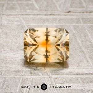 A 1.10-carat pale yellow-orange particolored Montana sapphire, cut in a rectangular faceted shape, showcased on a textured metallic surface with the “Earth’s Treasury” logo beneath it.