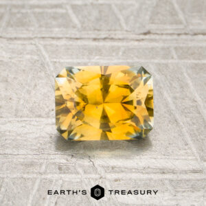 A radiant-cut, rectangular golden yellow Montana sapphire (0.98 carats, heated) is displayed on a textured gray surface, with the “Earth’s Treasury” logo and text at the bottom of the image.