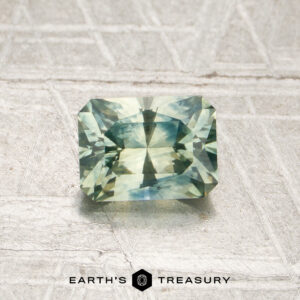 A 0.96-carat, rectangular, faceted mint green Montana sapphire (heated) is displayed on a textured pale background, with the words “EARTH’S TREASURY” beneath it.