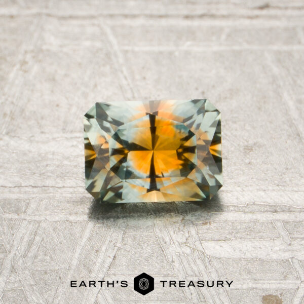 0.85-Carat Aqua-Orange Particolored Montana Sapphire (Heated) A 0.85-carat aqua-orange particolored Montana sapphire (heated) with a rectangular faceted cut is displayed on a textured surface above the Earth’s Treasury logo.
