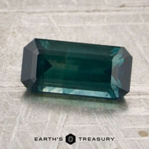A rectangular, emerald-cut 2.38-carat deep blue-green heated Montana sapphire rests on a textured light gray surface, with the logo and text “EARTH’S TREASURY” at the bottom.