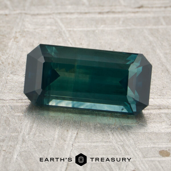 2.38-Carat Deep Blue-Green Montana Sapphire (Heated) A rectangular, emerald-cut 2.38-carat deep blue-green heated Montana sapphire rests on a textured light gray surface, with the logo and text “EARTH’S TREASURY” at the bottom.