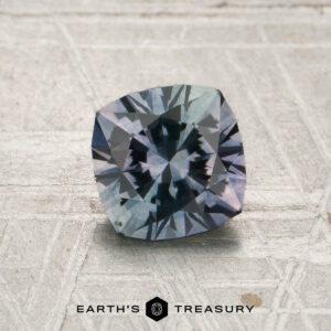 A 1.46-carat lilac-teal particolored Montana sapphire rests on a textured surface, with the “Earth’s Treasury” logo displayed at the bottom center.