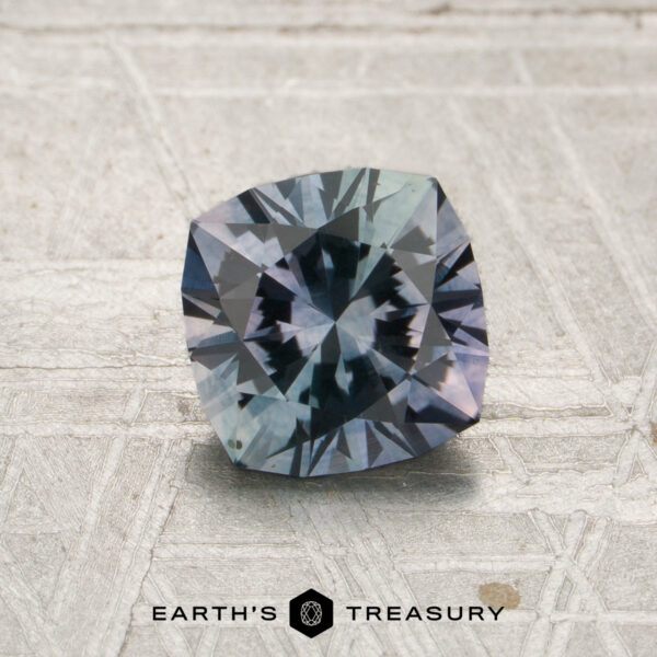 1.46-Carat Lilac-Teal Particolored Montana Sapphire (Heated) A 1.46-carat lilac-teal particolored Montana sapphire rests on a textured surface, with the “Earth’s Treasury” logo displayed at the bottom center.