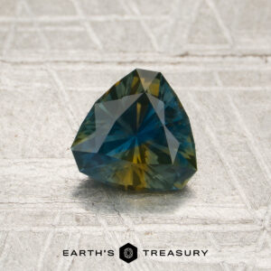 A 1.05-carat blue-yellow particolored Montana sapphire (heated), expertly faceted in a trilliant cut, is showcased on a textured surface. The “EARTH’S TREASURY” logo and text are visible at the bottom of the image.