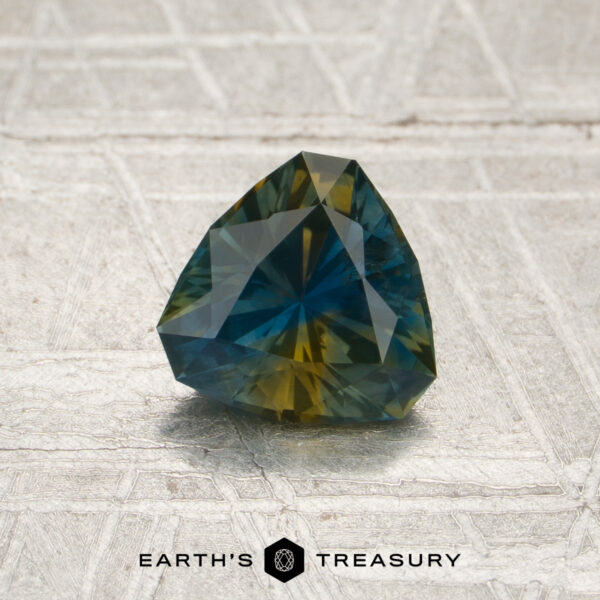 1.05-Carat Blue-Yellow Particolored Montana Sapphire (Heated) A 1.05-carat blue-yellow particolored Montana sapphire (heated), expertly faceted in a trilliant cut, is showcased on a textured surface. The “EARTH’S TREASURY” logo and text are visible at the bottom of the image.