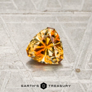 A 0.78-carat orange-light yellow particolored Montana sapphire, trillion-cut, rests on a textured gray surface, with the text “Earth’s Treasury” and a logo displayed below.