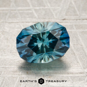 A 2.35-carat rich teal-blue particolored Montana sapphire (heated) rests on a textured surface with the Earth’s Treasury logo and text visible at the bottom.