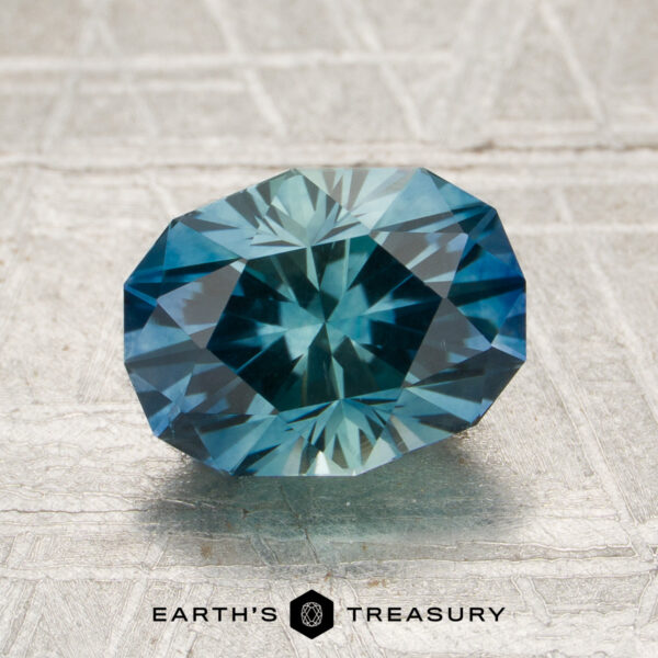 2.35-Carat Rich Teal-Blue Particolored Montana Sapphire (Heated) A 2.35-carat rich teal-blue particolored Montana sapphire (heated) rests on a textured surface with the Earth’s Treasury logo and text visible at the bottom.