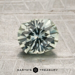 A 1.83-carat pale mint green Montana sapphire (heated), cut in a round faceted shape, rests on a textured gray surface above the “Earth’s Treasury” text and logo.