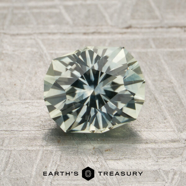1.83-Carat Pale Mint Green Montana Sapphire (Heated) A 1.83-carat pale mint green Montana sapphire (heated), cut in a round faceted shape, rests on a textured gray surface above the “Earth’s Treasury” text and logo.