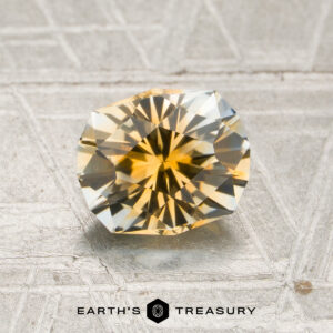 A 1.50-carat golden yellow-colorless particolored Montana sapphire is displayed on a textured surface, with the text “EARTH’S TREASURY” and a hexagon logo at the bottom.