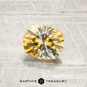 A 1.21-carat yellow-colorless particolored Montana sapphire (heated) rests on a textured, light-colored surface. The logo and text “Earth’s Treasury” appear at the bottom of the image.