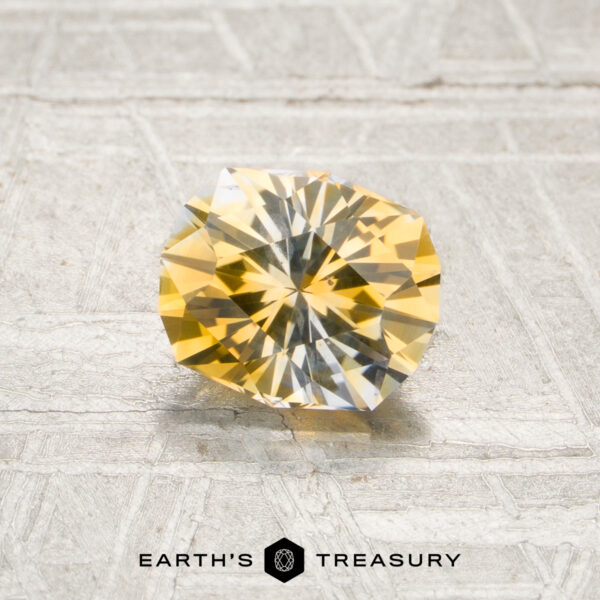 1.21-Carat Yellow-Colorless Particolored Montana Sapphire (Heated) A 1.21-carat yellow-colorless particolored Montana sapphire (heated) rests on a textured, light-colored surface. The logo and text “Earth’s Treasury” appear at the bottom of the image.