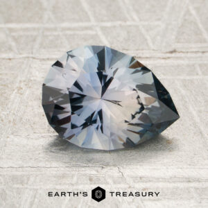 A 1.89-carat blue-gray Montana sapphire (heated) rests on a textured surface, with the Earth’s Treasury logo displayed at the bottom of the image.