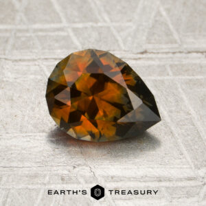 A 1.47-carat burnt orange Montana sapphire (heated), cut into a pear shape, rests on a textured surface, with “EARTH’S TREASURY” and a hexagonal logo displayed below.
