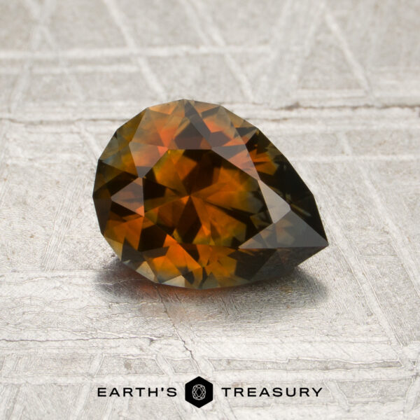 1.47-Carat Burnt Orange Montana Sapphire (Heated) A 1.47-carat burnt orange Montana sapphire (heated), cut into a pear shape, rests on a textured surface, with “EARTH’S TREASURY” and a hexagonal logo displayed below.