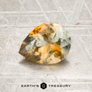 A faceted, pear-shaped 1.38-carat Orange-Near Colorless Particolored Montana Sapphire gemstone rests on a textured surface. The “Earth’s Treasury” logo and text appear at the bottom.