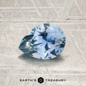 A 1.17-carat, pear-shaped, faceted Sky Blue Montana Sapphire (heated) is displayed against a textured, metallic surface with the text “EARTH’S TREASURY” at the bottom.