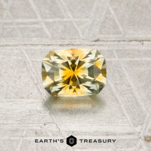 A 0.91-carat pale yellow-orange particolored Montana sapphire is displayed on a textured grey surface with the “EARTH’S TREASURY” logo at the bottom.