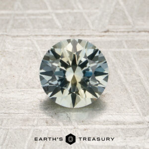 A 1.34-carat heated Montana sapphire, round and faceted with pale blue and yellow hues, is displayed on a textured light surface. The text “EARTH’S TREASURY” appears at the bottom of the image.
