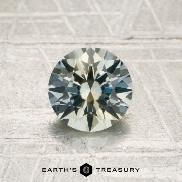 1.34-Carat Pale Blue-Yellow Particolored Montana Sapphire (Heated) A 1.34-carat heated Montana sapphire, round and faceted with pale blue and yellow hues, is displayed on a textured light surface. The text “EARTH’S TREASURY” appears at the bottom of the image.