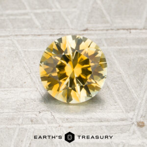A 1.17-carat yellow Montana sapphire (heated) with a brilliant cut is displayed on a textured gray surface, with the “Earth’s Treasury” logo and text visible at the bottom of the image.