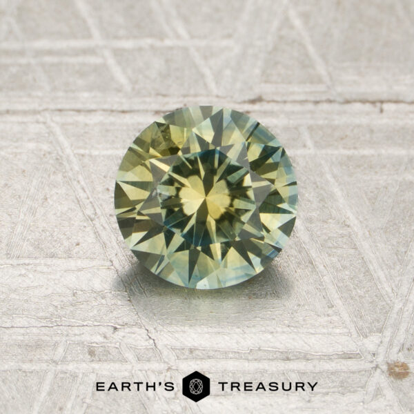 1.17-Carat Light Yellow-Blue Particolored Montana Sapphire (Heated) A round, faceted 1.17-carat light yellow-blue particolored Montana sapphire (heated) is displayed on a textured metallic surface with the “EARTH’S TREASURY” logo at the bottom.