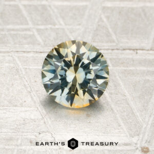 A 1.05-carat pale blue-yellow particolored Montana sapphire (heated), featuring a round brilliant cut, is displayed on a textured gray surface. The words “EARTH’S TREASURY” are visible at the bottom of the image.