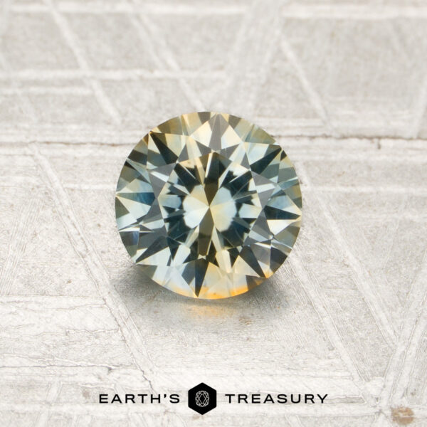 1.05-Carat Pale Blue-Yellow Particolored Montana Sapphire (Heated) A 1.05-carat pale blue-yellow particolored Montana sapphire (heated), featuring a round brilliant cut, is displayed on a textured gray surface. The words “EARTH’S TREASURY” are visible at the bottom of the image.