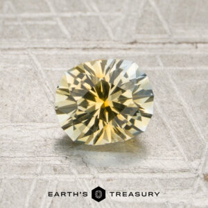 A 1.27-carat light yellow-colorless particolored Montana sapphire, expertly faceted in a round shape, is displayed on a textured metallic surface with the words “EARTH’S TREASURY” at the bottom.