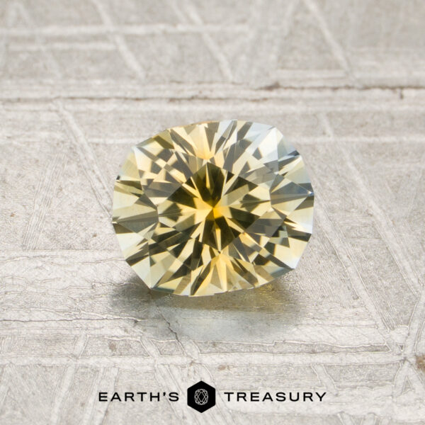 1.27-Carat Light Yellow-Colorless Particolored Montana Sapphire (Heated) A 1.27-carat light yellow-colorless particolored Montana sapphire, expertly faceted in a round shape, is displayed on a textured metallic surface with the words “EARTH’S TREASURY” at the bottom.