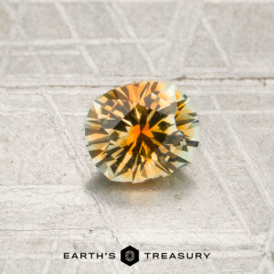 A faceted 0.84-carat orange-colorless particolored Montana sapphire with yellow, orange, and green tones rests on a textured silvery surface. The “Earth’s Treasury” logo and text appear at the bottom of the image.