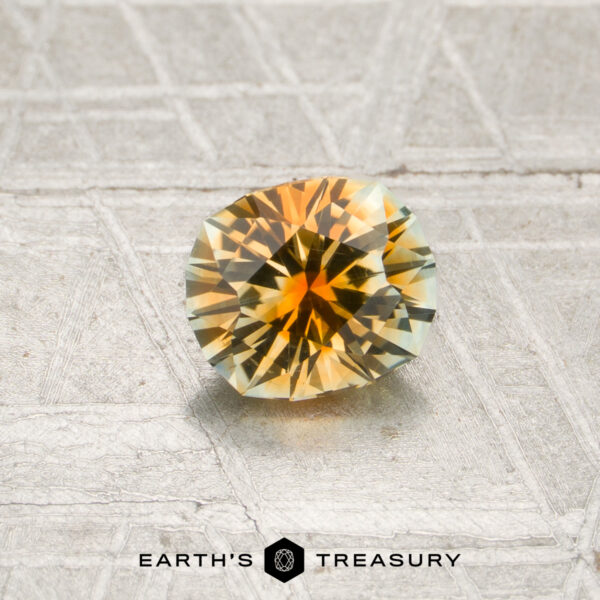 0.84-Carat Orange-Colorless Particolored Montana Sapphire (Heated) A faceted 0.84-carat orange-colorless particolored Montana sapphire with yellow, orange, and green tones rests on a textured silvery surface. The “Earth’s Treasury” logo and text appear at the bottom of the image.