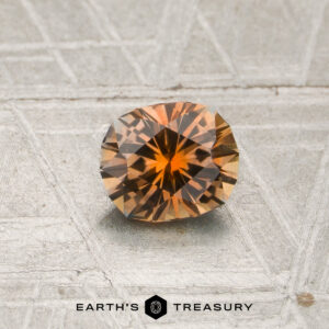 A 0.83-carat round, faceted orange Montana sapphire (heated) is displayed on a textured, light gray surface with the “Earth’s Treasury” text and logo at the bottom.