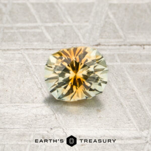 A round, faceted 0.80-carat orange-near colorless particolored Montana sapphire rests on a textured metallic surface above the words “Earth’s Treasury.”.