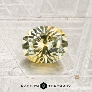 A 1.40-carat light yellow Montana sapphire (heated), round and faceted, is displayed on a textured, light-colored surface with the “EARTH’S TREASURY” logo beneath it.