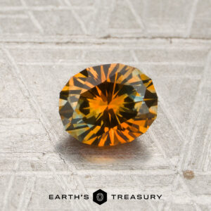 A 1.09-carat rich orange-aqua particolored Montana sapphire (heated), faceted in a round cut, displays its vibrant golden-orange tones atop a textured gray surface, with the “EARTH’S TREASURY” branding and logo featured at the bottom.