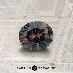 A 1.05-carat tricolor Montana sapphire (heated) with blue and reddish hues rests on a textured metallic surface; the “EARTH’S TREASURY” logo appears at the bottom.