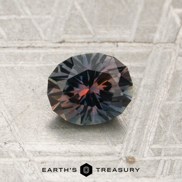 1.05-Carat Tricolor Montana Sapphire (Heated) A 1.05-carat tricolor Montana sapphire (heated) with blue and reddish hues rests on a textured metallic surface; the “EARTH’S TREASURY” logo appears at the bottom.