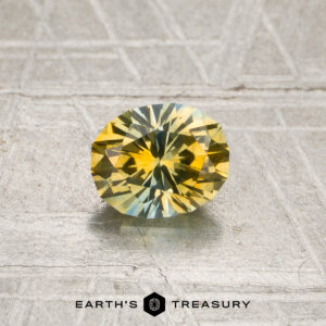 A faceted oval 0.88-carat yellow-sky blue particolored Montana sapphire (heated) rests on a textured surface, with the text “EARTH’S TREASURY” and a geometric logo displayed below it.