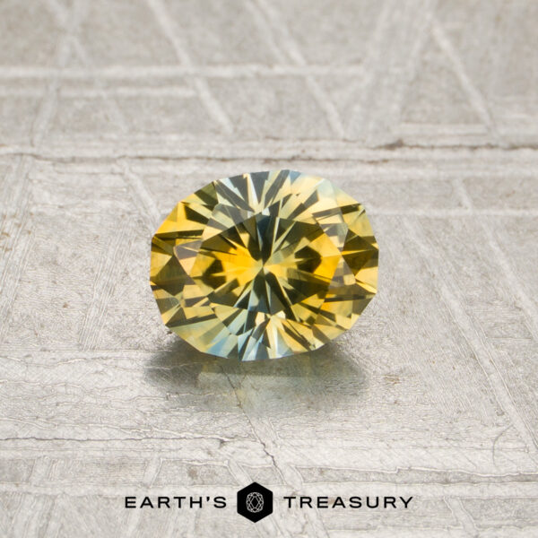 0.88-Carat Yellow-Sky Blue Particolored Montana Sapphire (Heated) A faceted oval 0.88-carat yellow-sky blue particolored Montana sapphire (heated) rests on a textured surface, with the text “EARTH’S TREASURY” and a geometric logo displayed below it.