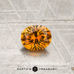 A 0.74-carat heated Montana sapphire, faceted in orange, rests on a textured gray surface, with the text “EARTH’S TREASURY” and a hexagonal logo displayed below.