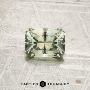 A rectangular, pale mint green faceted gemstone—identified as a 0.97-carat Mint Green Montana Sapphire (Heated)—is displayed on a light textured surface. The logo and text “EARTH’S TREASURY” appear at the bottom of the image.
