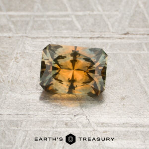 A rectangular, multicolored 0.96-carat orange-teal particolored Montana sapphire (heated) rests on a textured, light surface, with the Earth’s Treasury logo visible at the bottom.