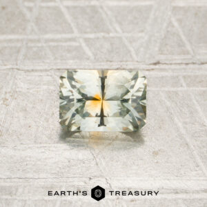 A 0.80-carat, rectangular, faceted Montana sapphire (heated) with light yellow and green hues is displayed on a textured, light-colored surface above the Earth’s Treasury logo.