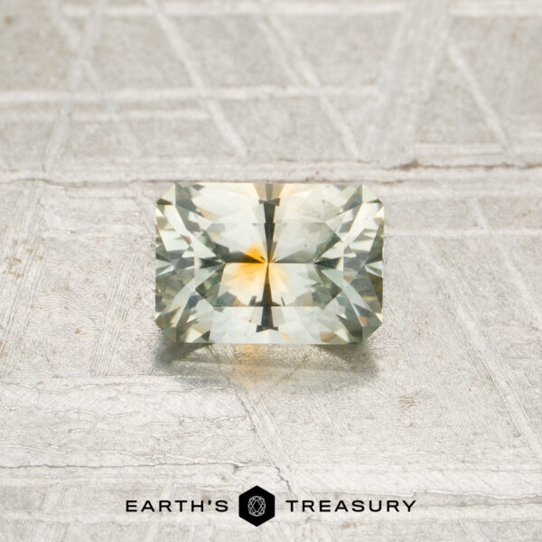 0.80-Carat Pale Mint Green-Orange Particolored Montana Sapphire (Heated) A 0.80-carat, rectangular, faceted Montana sapphire (heated) with light yellow and green hues is displayed on a textured, light-colored surface above the Earth’s Treasury logo.