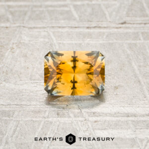 A 0.78-carat orange Montana sapphire (heated), cut in a rectangular shape with faceted edges, is displayed on a textured surface with the “EARTH’S TREASURY” logo at the bottom.