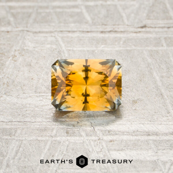 0.78-Carat Orange Montana Sapphire (Heated) A 0.78-carat orange Montana sapphire (heated), cut in a rectangular shape with faceted edges, is displayed on a textured surface with the “EARTH’S TREASURY” logo at the bottom.