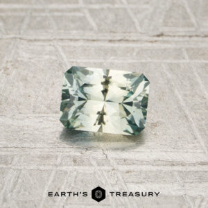 A 0.77-carat, rectangular faceted Montana sapphire with a pale green hue is displayed on a textured, light-colored surface. The words “EARTH’S TREASURY” appear at the bottom.